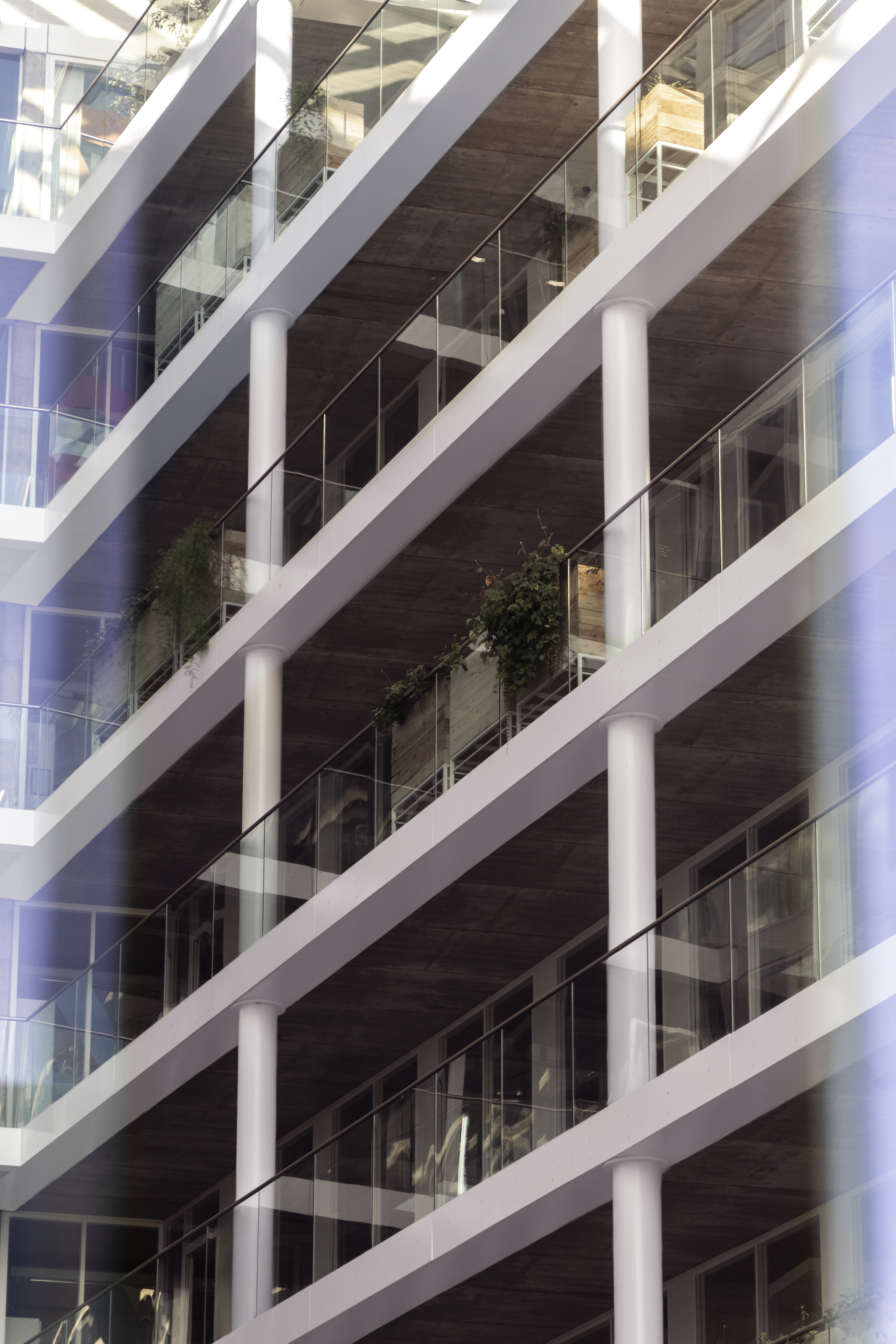 La photographie présente un bâtiment moderne aux lignes géométriques épurées. L'image met en avant plusieurs étages de balcons successifs, soutenus par des colonnes blanches. Chaque balcon est délimité par des garde-corps en verre, renforçant l'impression de transparence et d'ouverture. Quelques touches de végétation, disposées dans des bacs, apportent une note de verdure à l'ensemble. L'image est structurée par des lignes horizontales et verticales qui attirent le regard vers les hauteurs du bâtiment.